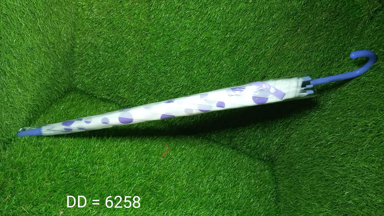 A closed white umbrella with purple geometric patterns and a blue handle lies on a green artificial grass surface.