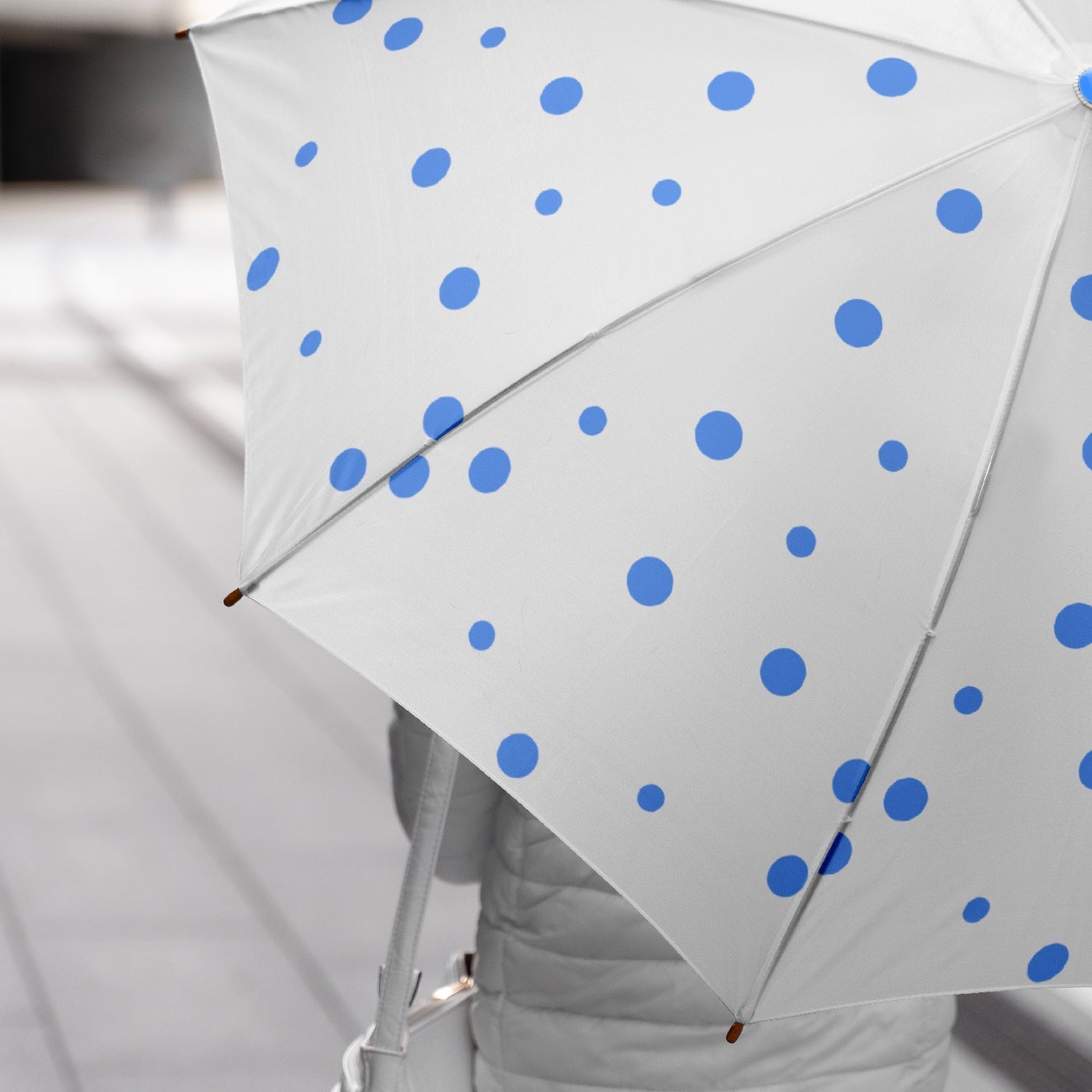 A person holds an open white umbrella with blue polka dots, with a gray background and a hint of a jacket.