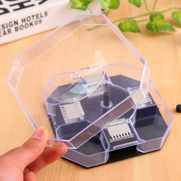 A hand lifts the clear plastic lid of a hexagonal trap, revealing the dark gray base and internal mechanisms on a wooden