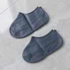 Two dark grey silicone shoe covers are laid flat on a light grey marble surface, showcasing their textured bottoms.