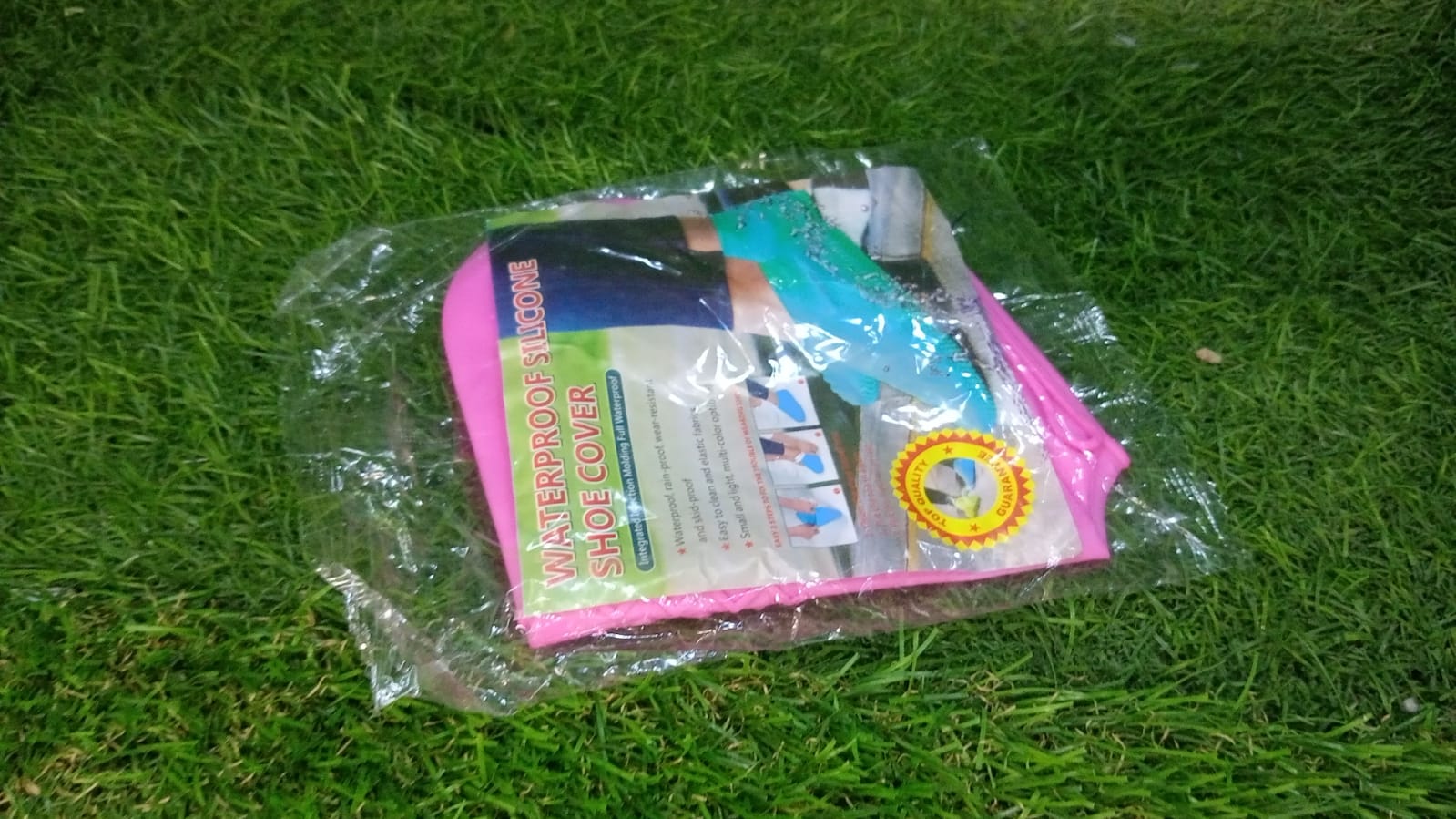 A clear plastic package containing pink and blue silicone shoe covers with product information, resting on green grass.